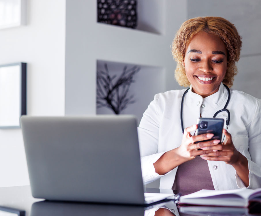 Clinician smiling at a smartphone at her desk, with a laptop open beside her